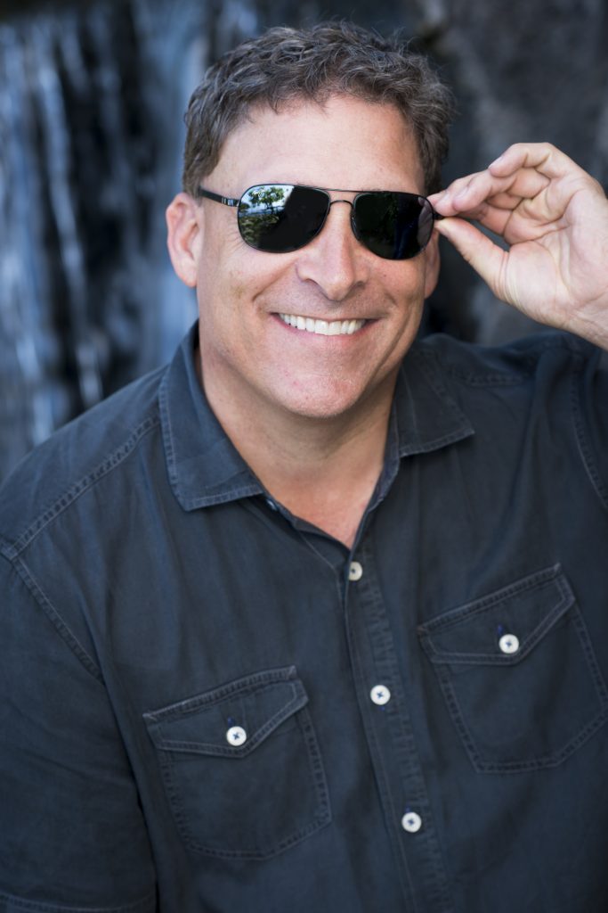 Man (Brett Hatch) in black button up and grey Maui Jim aviator sunglasses poses in front of a blurred grey background
