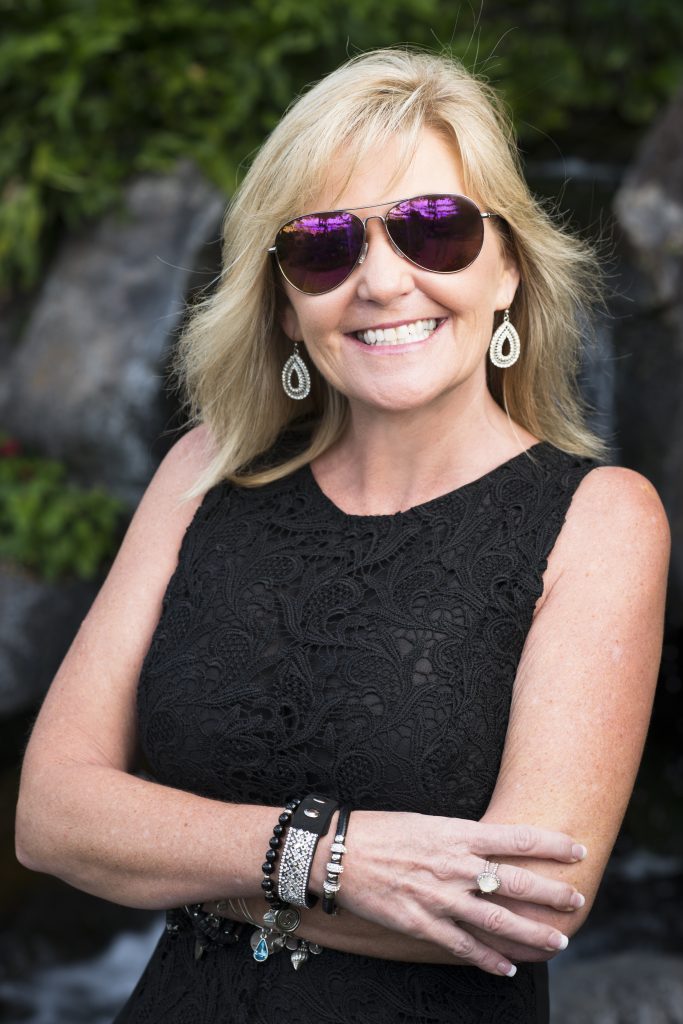 Karen Suttle - blonde woman wearing a black tank top and Maverick Maui Jims with pink mirror lenses. She poses with arms folded in front of a blurred background.