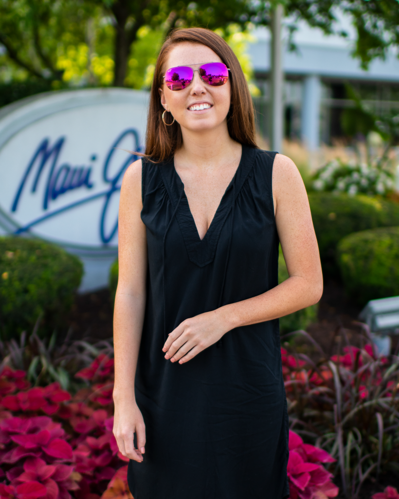 Woman in black dress and pink sunglasses poses in front of the Maui Jim sign