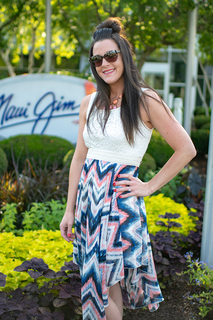 Woman (Devon Thompson) in brown sunglasses, white tank top, and multicolor skirt poses in front of the Maui Jim sign