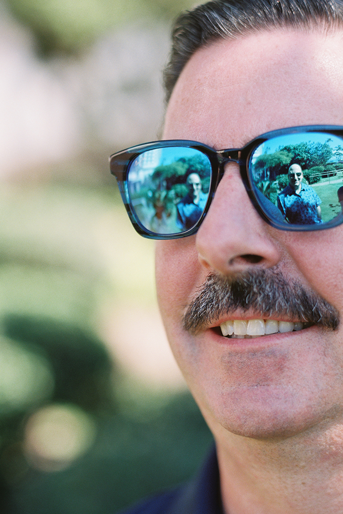Close up of a brunette man with a mustache (Jonathan Richards) wearing Maui Jim sunglasses with a black and blue striped frame and blue lenses.